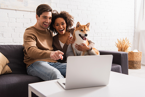 Positive man pointing at blurred laptop near african american girlfriend with shiba inu at home