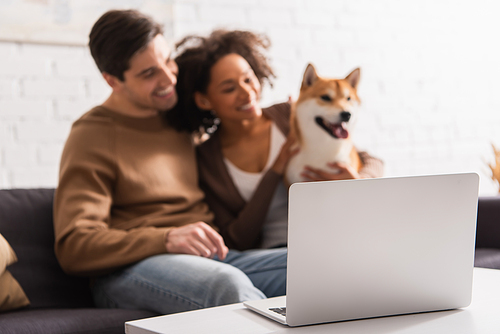 Laptop on coffee table near blurred multiethnic couple with shiba inu dog at home