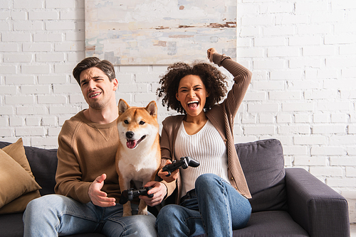 KYIV, UKRAINE - DECEMBER 22, 2021: Cheerful african american woman playing video game with upset boyfriend near shiba inu at home