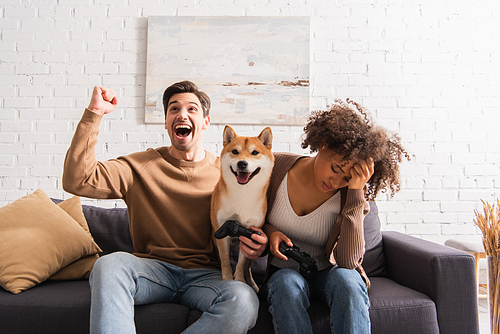 KYIV, UKRAINE - DECEMBER 22, 2021: Excited man holding joystick near sad african american girlfriend and shiba inu at home
