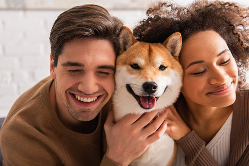 Cheerful man with closed eyes petting shiba inu near african american girlfriend at home