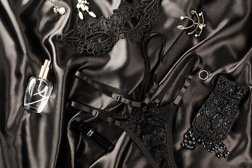 Top view of sexual mask near panties and perfume on black satin background