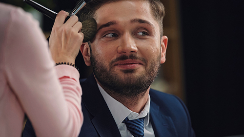 blurred makeup artist applying face powder on smiling man in formal wear