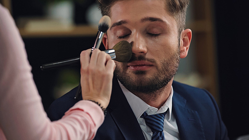 blurred woman doing makeup to young man with closed eyes