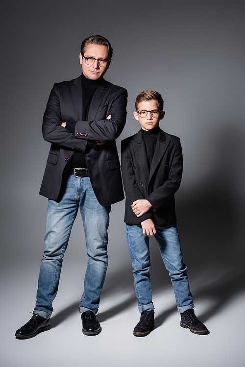 Full length of stylish boy in jacket and jeans standing near father on grey