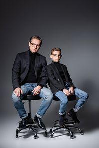 Stylish father and preteen son sitting on chairs on grey