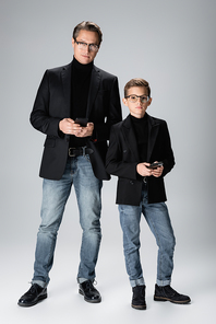 Full length of stylish parent and boy holding mobile phones on grey background