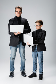 Full length of trendy man and boy using laptops on grey background