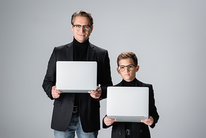 Stylish preteen son holding laptop near dad at home isolated on grey