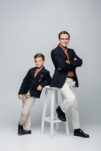 Positive and stylish father and son posing near chair on grey background