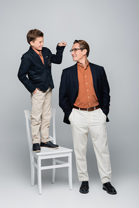 Smiling boy pointing at stylish dad while standing on chair on grey background