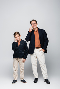 Cheerful stylish father and son talking on smartphones on grey background