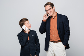 Smiling father and son in jackets talking on smartphones isolated on grey