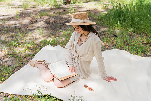 smiling young woman in straw hat reading book in forest