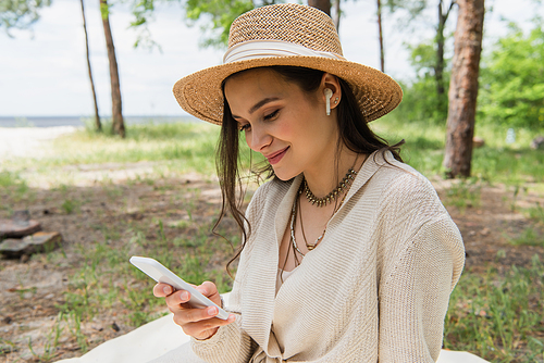 positive young woman in straw hat and wireless earphones using smartphone in forest