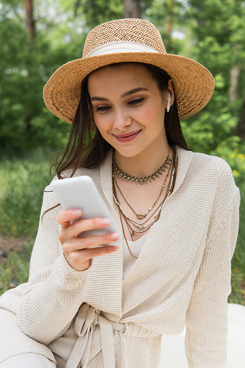 happy young woman in straw hat and wireless earphones using blurred smartphone in forest