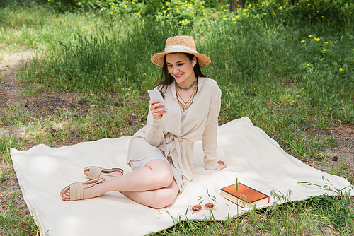 smiling young woman in straw hat and wireless earphones using smartphone while sitting on picnic blanket