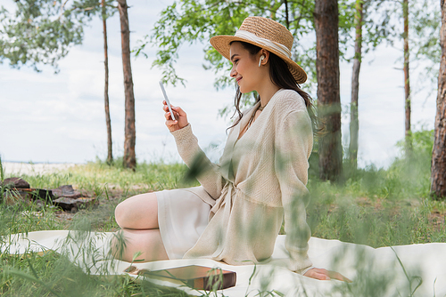 smiling woman in straw hat and wireless earphones using smartphone while sitting on picnic blanket