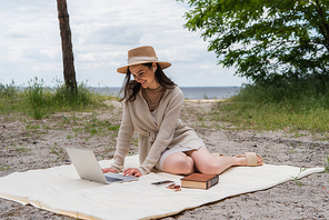 happy woman in straw hat and earphones using laptop while sitting on picnic blanket near smartphone, sunglasses and book