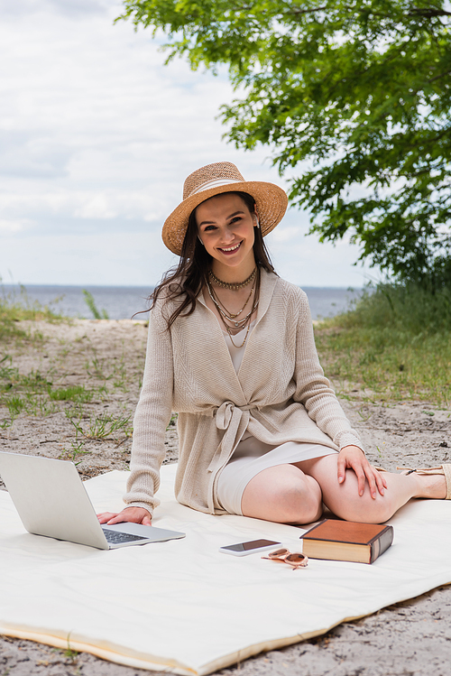 cheerful woman in straw hat and earphones using laptop while sitting on picnic blanket near smartphone, sunglasses and book