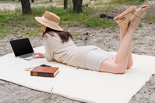 woman in straw hat and earphones using laptop with blank screen while lying on picnic blanket near smartphone, sunglasses and book