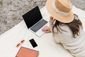 high angle view of woman in straw hat using laptop with blank screen while lying on picnic blanket near smartphone, sunglasses and book