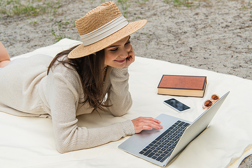joyful freelancer in straw hat using laptop while lying on picnic blanket near smartphone, sunglasses and book