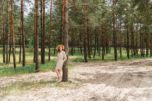 full length of cheerful young woman in straw hat standing in woods