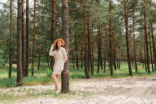 full length of cheerful young woman adjusting straw hat and standing in woods