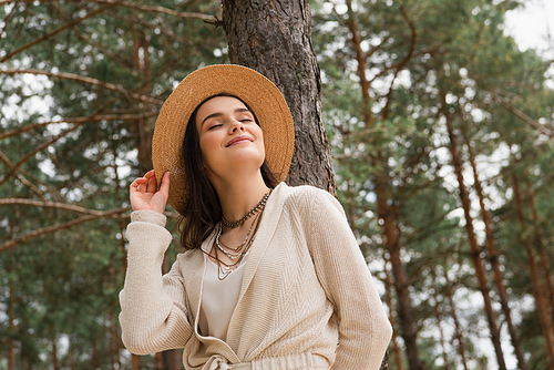 cheerful young woman adjusting straw hat and smiling in woods