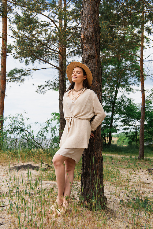 full length of smiling young woman in straw hat standing in forest