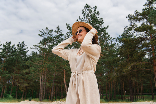 cheerful young woman in sunglasses adjusting straw hat near forest