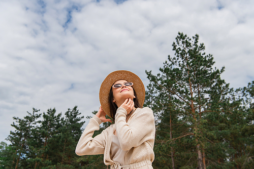 low angle view of cheerful young woman in sunglasses and straw hat looking up near forest