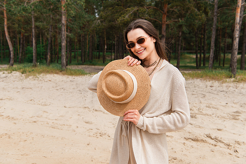 cheerful young woman in sunglasses holding straw hat near green forest