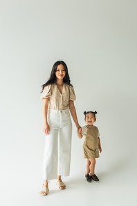 full length of stylish asian mother holding hands with happy toddler daughter on grey