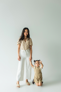 full length of stylish smiling asian mother holding hands with toddler daughter on grey