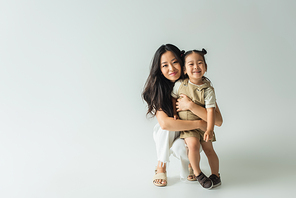full length of stylish asian mother hugging toddler daughter on grey