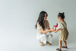 full length of stylish asian mother receiving present from toddler daughter on grey