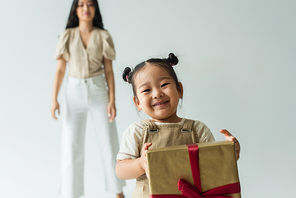 happy asian toddler girl holding present near blurred mother isolated on grey