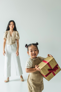 happy asian toddler kid holding present near blurred mother on grey