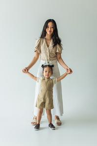 full length of happy asian toddler kid and stylish mother holding hands on grey