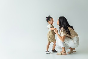 full length of happy asian toddler kid holding hands with stylish mother on grey