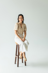 full length of asian brunette woman in trendy outfit sitting on chair on grey