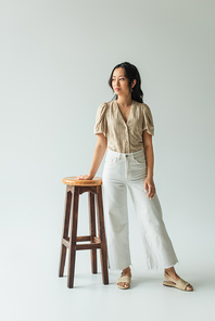 full length of asian brunette woman in trendy outfit leaning on wooden chair on grey