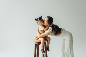happy asian toddler girl sitting on chair and hugging mother isolated on grey