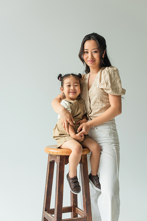 asian toddler girl sitting on chair and hugging with mother isolated on grey