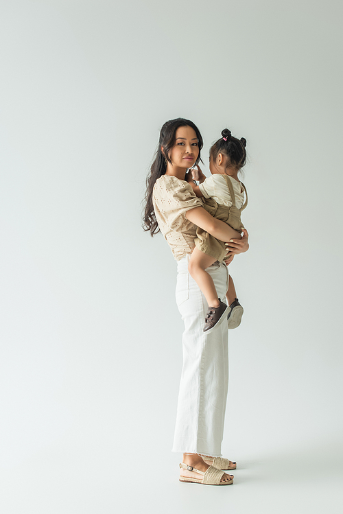 full length of happy asian woman holding in arms toddler daughter on grey