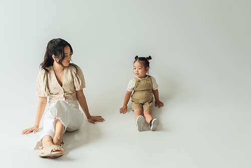 full length of asian mother sitting and looking at toddler daughter on grey