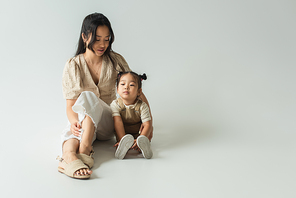 full length of asian mother sitting with toddler daughter on grey