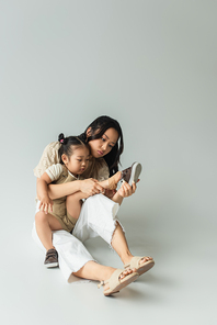 full length of asian mother sitting and wearing shoe of foot of toddler daughter on grey